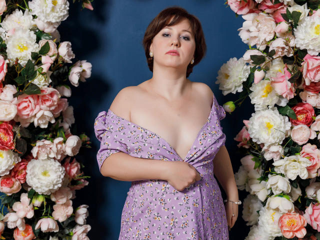 MirandaBrooks with short brown hair, pale skin, and small tits, wearing a lilac dress, off-shoulder, revealing cleavage, standing between two floral walls, looking up.