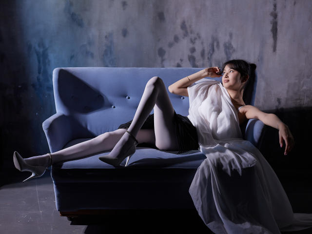 Sheila, peau pâle, cheveux noirs, chemisier blanc, short noir, collants gris, talons blancs, allongée sur un canapé bleu, bras droit levé, jambe gauche tendue, robe blanche drapée..