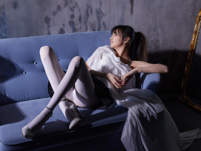 Sheila: Sentada em um sofá azul, vestindo um vestido branco, calcinha preta, meias cor de rosa claro, saltos brancos, braços relaxados, pernas cruzadas, rabo de cavalo, parede cinza, espelho dourado, luz suave, pose relaxada..