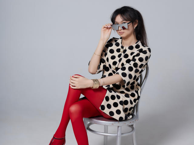 SheilaPass sitting on a chair, wearing a black and white polka dot dress, red tights, and black shoes, adjusting her glasses, with a bracelet on her wrist.
