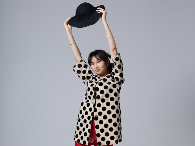 SheilaPass holding a black hat above her head with both arms, wearing a polka-dotted beige shirt and red pants, looking slightly to the side with a subtle smile.