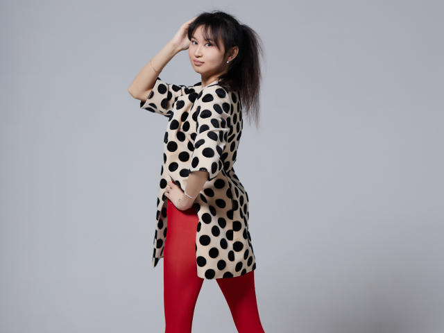 SheilaPass with black polka dot shirt, red tights, and messy black hair, standing sideways, hand in hair, looking over shoulder, gray background.