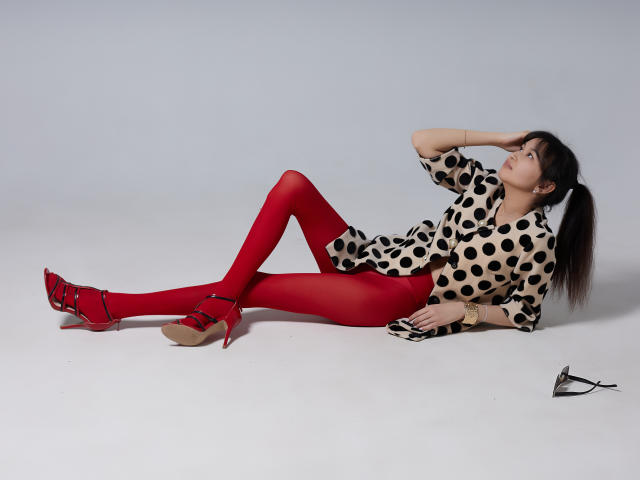 SheilaPass with red tights, polka dot shirt, and red heels, lying on white floor, hand on head, black ponytail, black and white shoes to the right.