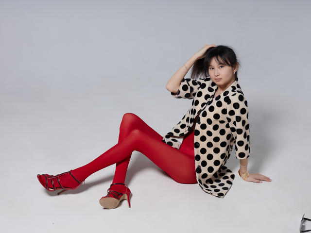 Sheila, con collant rossi, camicia a pois, tacchi rossi e biancheria intima rossa, seduta sul pavimento bianco, con una mano tra i capelli, che guarda la fotocamera. Seno, sedere, vagina, carina, attraente..