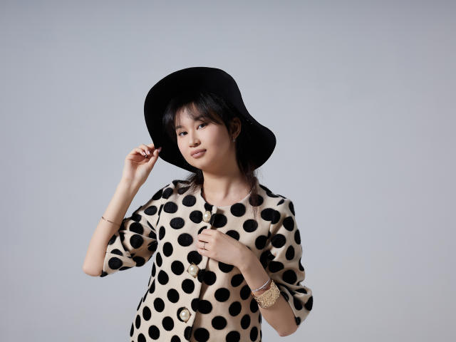 SheilaPass has black polka dot dress, black hat, and gold bracelets, adjusting hat with right hand, looking slightly to the side, with a subtle, confident smile.