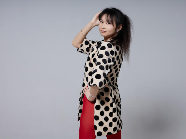 SheilaPass met een zwart topje met stippen, rode leggings, staand met de zijkant naar de camera, hand in het haar, kijkt over de schouder, klein achterwerk, gemiddelde borsten, lichte huid, Aziatisch, steil zwart haar, grijze achtergrond..