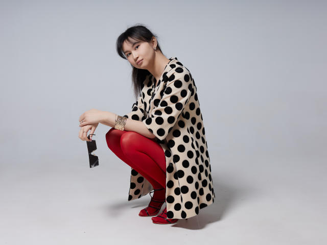 SheilaPass está agachada, vestindo um vestido bege com bolinhas, meias vermelhas e saltos vermelhos, segurando óculos de sol pretos, com cabelo preto liso e um sorriso discreto, parecendo adorável..