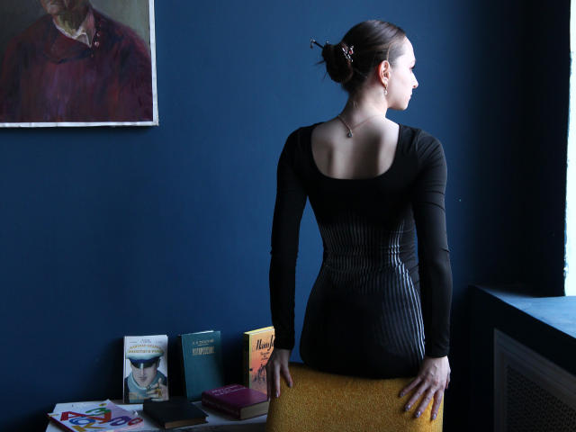 JasmineNeal stands with back to camera, black dress, bare shoulders, brown bun, hands on yellow chair, blue wall, portrait above, books and photo on left.