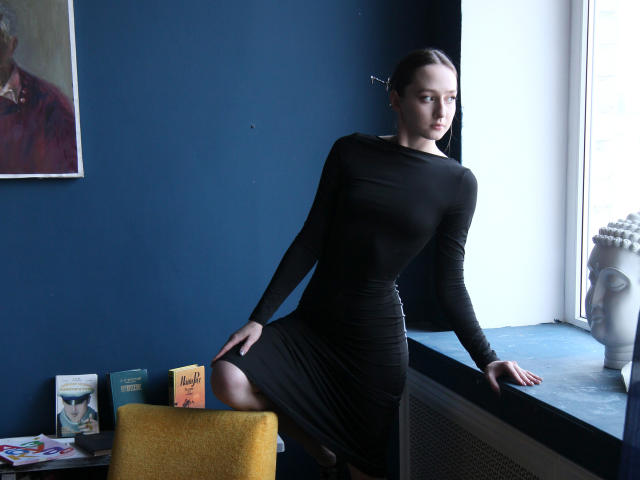 JasmineNeal in a tight black dress, standing with one leg raised, showing her ass, by a window with blue walls, yellow chair, and books. Her tits are small.