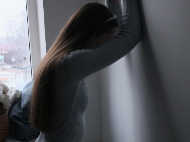 JasmineNeal with long brown hair, wearing a gray rib-knit sweater, arms up against a white wall, head down, in a dimly lit room with a window.