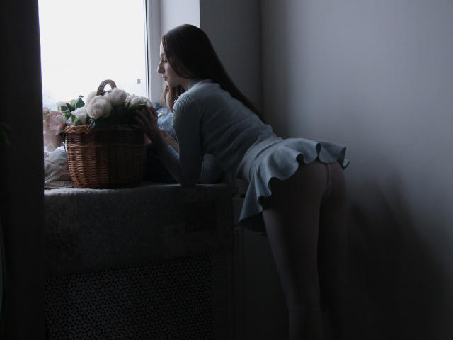JasmineNeal leans on a window, wearing a light blue, ruffled, short dress, exposing her ass, with long brown hair, looking outside, next to a wicker basket of roses.