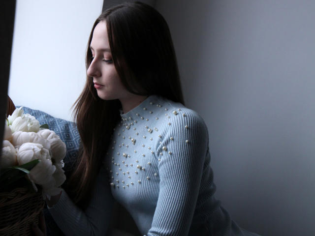 JasmineNeal with long brown hair, wearing a light blue, ribbed, studded top, holding a bouquet of white peonies, looking down, sitting near a window, in soft light.