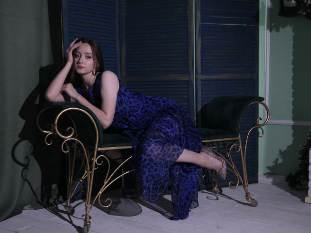 JasmineNeal, with pale skin and brown hair, leans on gold metal chairs wearing a blue leopard-print dress, showing her ass, in a dimly lit room.