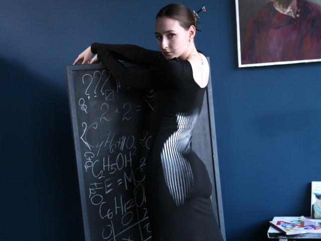 JasmineNeal with black dress, off-shoulder, standing by chalkboard, looking over shoulder, dark hair in bun, blue wall, framed picture, small tattoo on right shoulder, blackboard numbers.