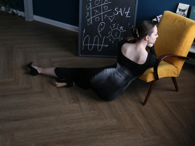 JasmineNeal is lying on her stomach in a black dress with her ass raised, wearing black high heels, and resting her head on a yellow chair.