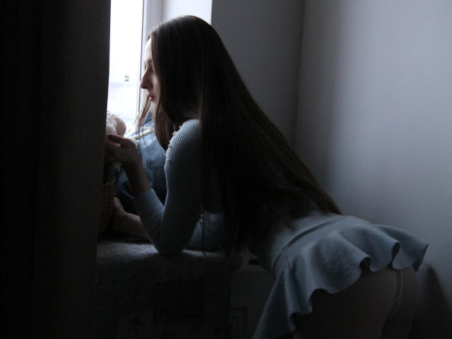 JasmineNeal with long brown hair, leaning on a windowsill, wearing a light blue dress with a flirty skirt, showing her white panties and ass, looking outside.
