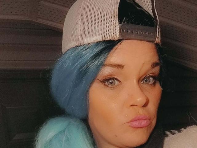 HotForYu with blue hair, wearing a backwards grey cap, has puffy pink lips and a neutral expression. Her ass is not visible, tits are not visible.