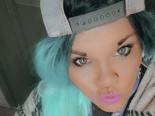 HotForYu with teal hair, wearing a grey cap, has a pouty face, purple lipstick, and green eyes. Her nose has a small stud, and she's looking up.