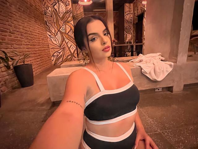 NovaDixon with dark hair, red lipstick, black and white sports bra, showing her tits, tattoo on arm, holding phone, standing in dimly lit room, white clothes on bench.