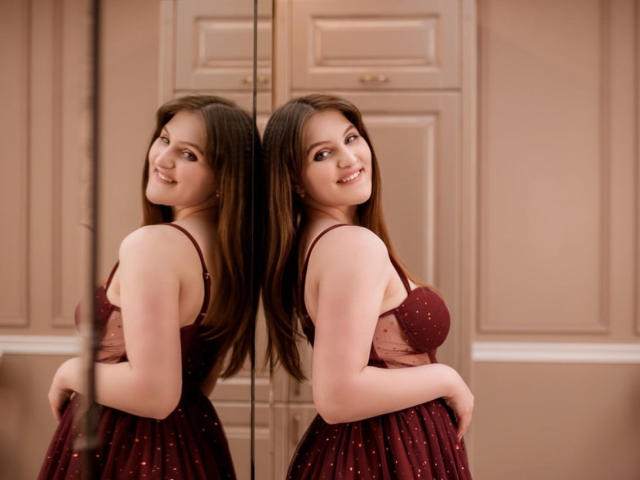 EmmyCharm with long brown hair, smiling at her reflection in a mirror. She's wearing a red, polka-dotted dress, showing her tits and looking cute. Mirror reflection included.