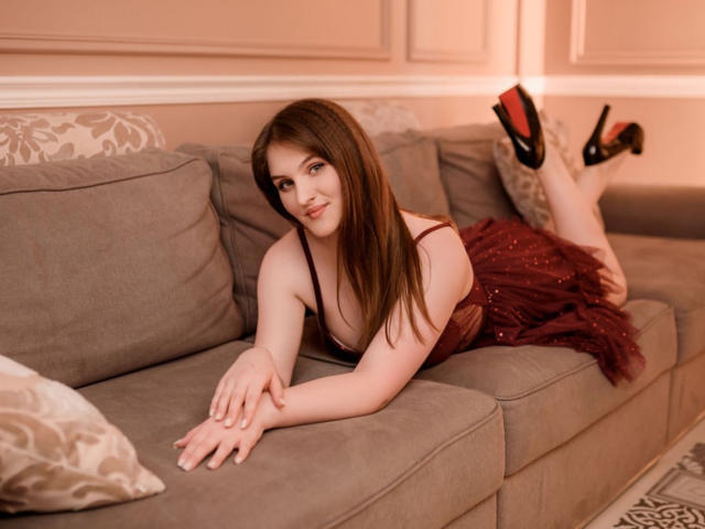 EmmyCharm lies on a grey couch, legs up, wearing a red, sparkly dress, black high heels, and a cute smile, showing her tits and ass. Brown hair, fair skin.