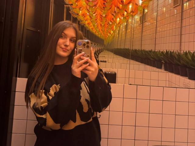 EmmyCharm with long brown hair, black and beige camouflage sweater, holding phone, smiling, standing in bathroom with orange flower decor above, white tiled walls, mirror reflection.