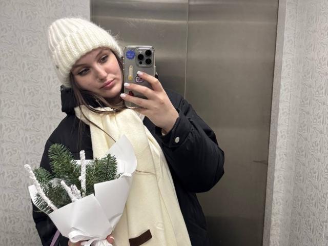 EmmyCharm with brown eyes, wearing a white knit hat, black coat, and cream scarf, holding a bouquet of pine, taking a mirror selfie in an elevator.