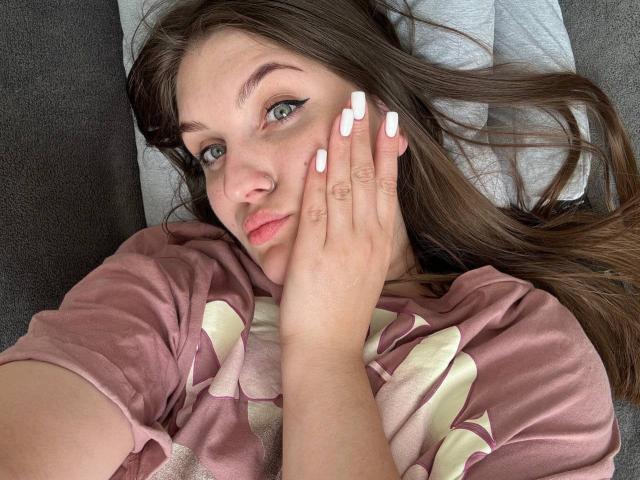 EmmyCharm with green eyes, brown hair, and white nails, touching her cheek with her hand, wearing a pink shirt with white floral patterns, lying on a grey surface.