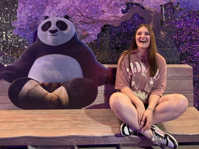 EmmyCharm, met een grote glimlach, zittend met gekruiste benen op een houten bankje, gekleed in een roze hoodie en witte korte broek, naast een levensgrote panda met een ontspannen, glimlachend gezicht..
