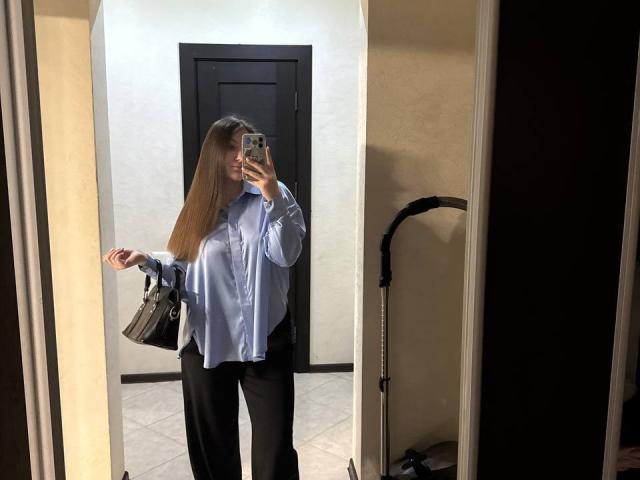EmmyCharm with long brown hair, wearing a blue shirt and black pants, holds a gray bag, standing in front of a mirror, taking a selfie with her phone.