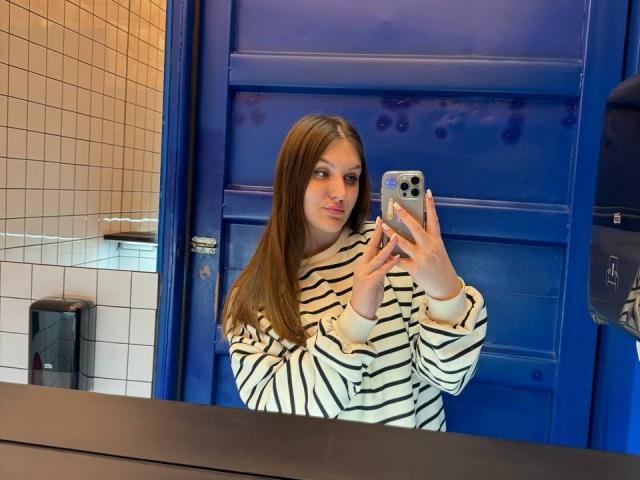 EmmyCharm met lang bruin haar, een gestreepte trui, die een telefoon vasthoudt en een spiegel-selfie maakt in een blauwe badkamer. Haar borsten zijn klein, haar achterkant is niet zichtbaar. Ze kijkt naar de camera..