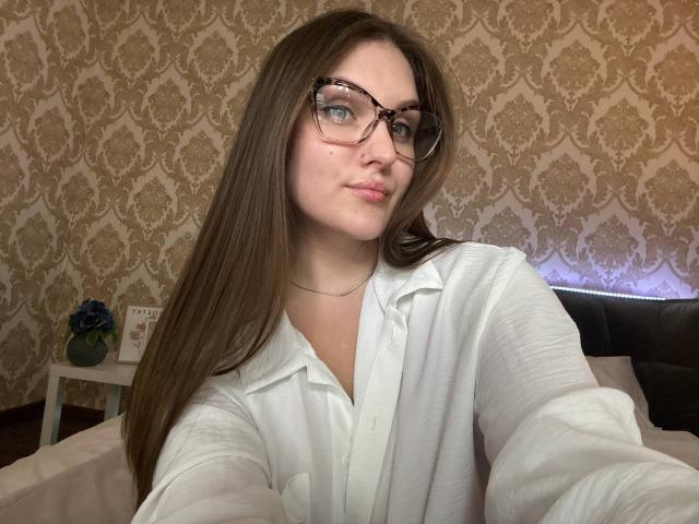EmmyCharm, con cabello castaño largo, gafas y una camisa blanca abotonada, tomando un selfie, mostrando una ligera sonrisa, en un dormitorio con papel tapiz estampado..