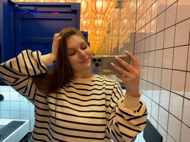 EmmyCharm with brown hair, striped shirt, holding phone in mirror, blue door, white tiled wall, smiling, hand in hair, phone case with purple sticker, cute.