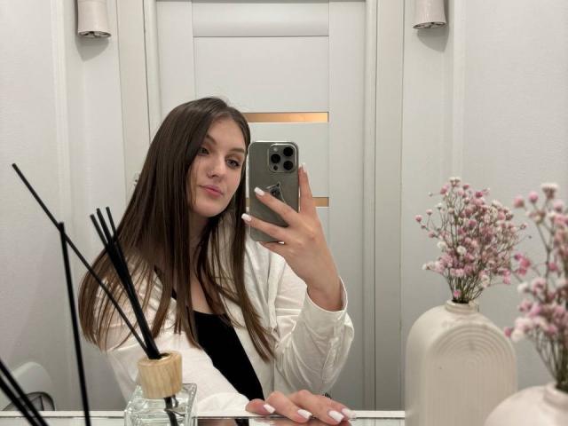 EmmyCharm with long brown hair, white shirt, black top, taking a mirror selfie with an iPhone, holding it in her right hand, showing her white nails.
