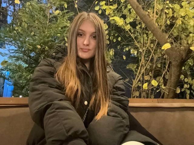 EmmyCharm with long brown hair, green puffy jacket, and small nose ring, sitting on brown bench, looking at camera, subtle smile.