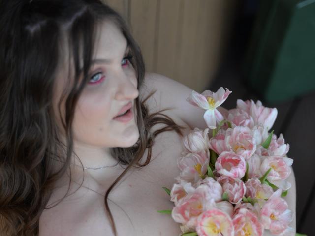 EmmyCharm with pink eyeliner, holding a bouquet of pink and white tulips, her bare tits visible, brown wavy hair, soft smile, silver necklace, slightly blurred background.