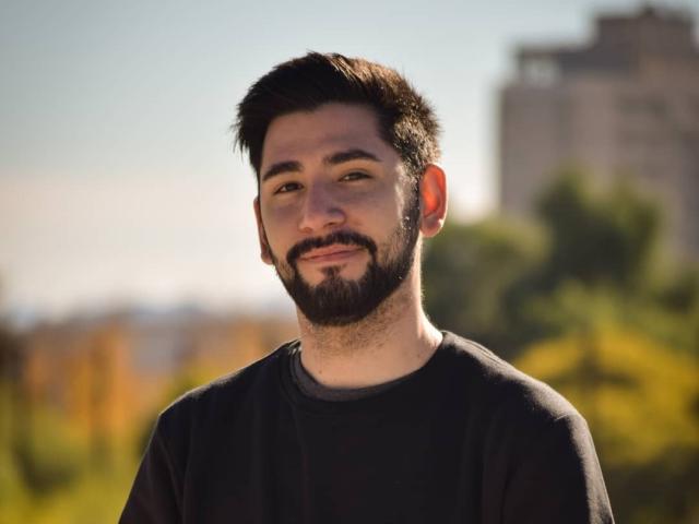 ElMagico has a dark beard and short, messy black hair. He's wearing a black shirt and smiling softly with a slight chin tuft. His skin is light.