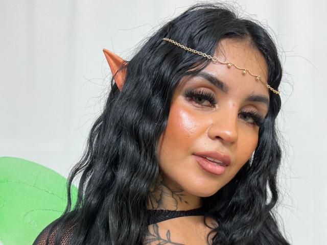 GabrielleKing with wavy black hair, brown skin, elf ears, gold chain headpiece, and a tattoo on her neck, showing off her shiny face and pouty lips.