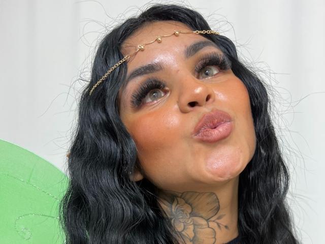 GabrielleKing, met zwart golvend haar, een gouden ketting als hoofdsieraad en volle lippen, laat haar grote tatoeage van een bloem op haar nek zien. Groene achtergrond..