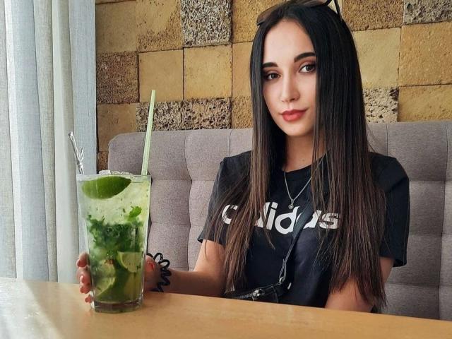 MissAnoliya-hot with long brown hair, wearing a black Adidas shirt, holding a green drink with a straw, sitting at a wooden table, looking at the camera.