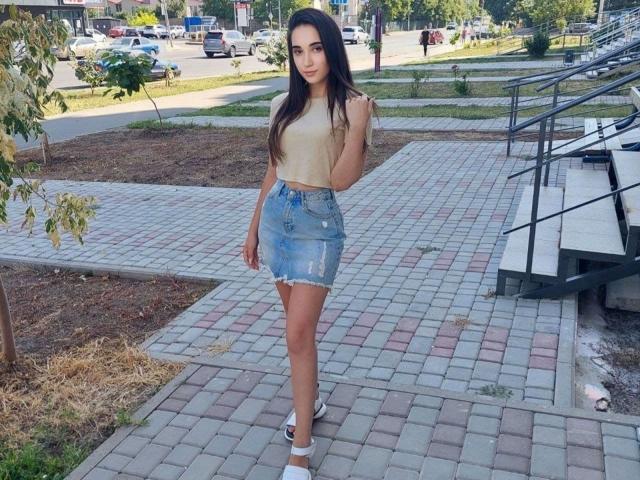 MissAnoliya-hot with long dark hair, tan sleeveless top, blue distressed denim skirt, white heels, standing on a brick pathway, hand near her face, sunny day.