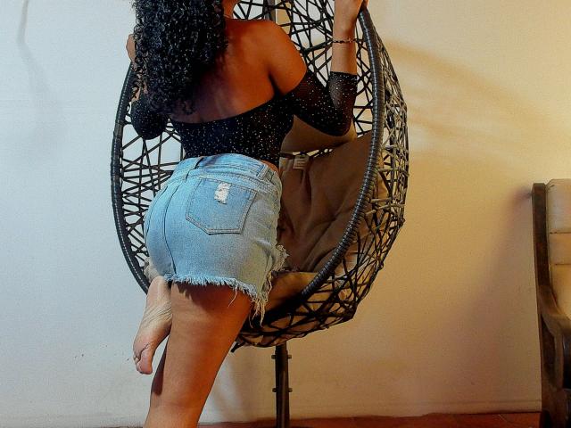 NatalyBlonder with curly black hair, off-shoulder black top, and frayed denim shorts, standing in a hanging chair, showing her ass and back, with one foot raised.