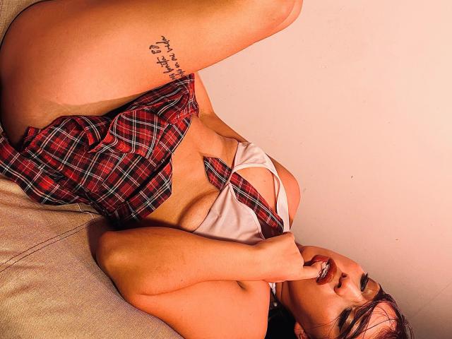 Violetta Roa, con grandes senos, vestida con una falda escocesa roja y una blusa blanca, acostada en un sofá, mordiéndose el dedo, mostrando un tatuaje en su muslo derecho..