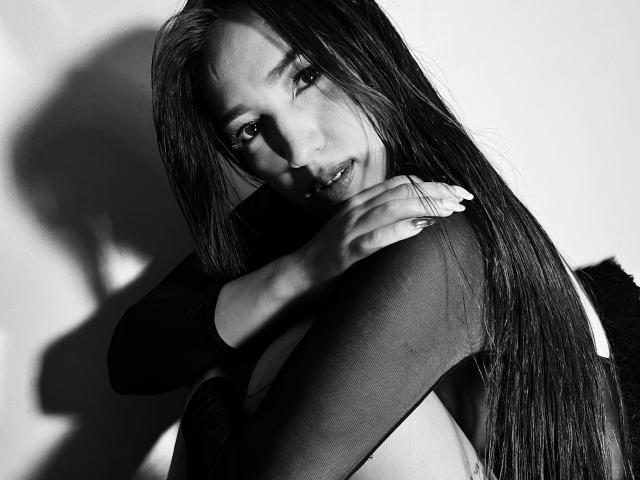 Fotografía en blanco y negro de ViolettaRoa con cabello largo y oscuro, vistiendo una blusa transparente, con los brazos cruzados sobre el pecho, mirando a la cámara con una sonrisa sutil, con una sombra en la pared..