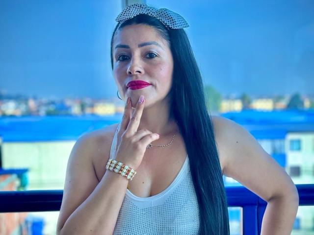 KinkyVane with long black hair, red lipstick, white mesh top, and checkered headband, touching finger to lips, wearing gold bracelet, blue background, looking sexy and confident.