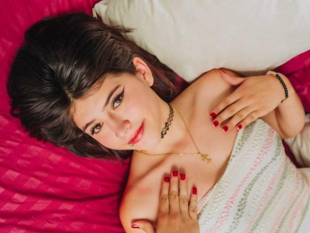 RachelHottie with dark hair, red nails, black choker, gold cross necklace, lying on pink bed, white pillow, hands on her tits, smiling, looking at camera.