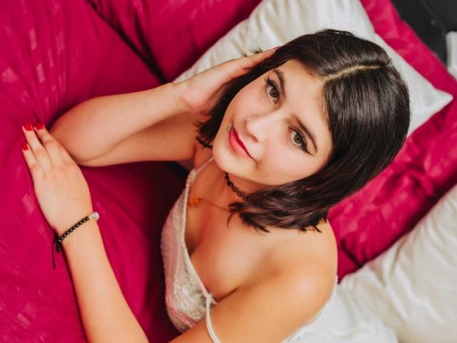 RachelHottie with short black hair, nose piercing, and red lipstick, lying on pink and white pillows, wearing a white lace top, looking up with a slight smile, red nails.
