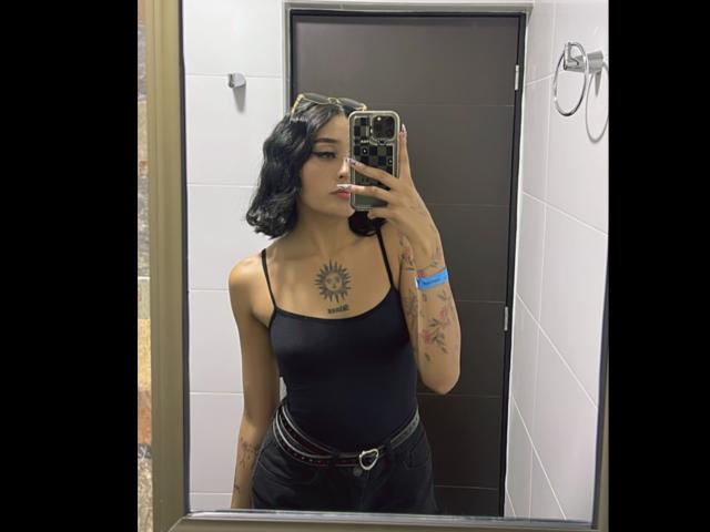 SalomeMille a les cheveux noirs et ondulés, un tatouage de soleil sur la poitrine, des tatouages sur les bras, elle porte un débardeur noir et un pantalon noir taille haute, et tient un téléphone devant un miroir..