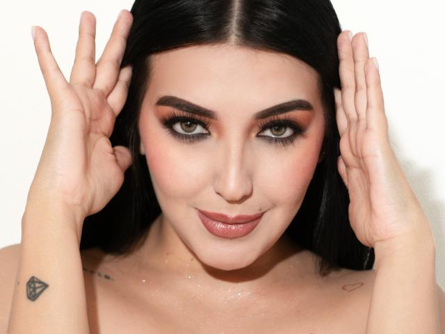 Ariana Cortes, com cabelo preto, olhos verdes e lábios rosados, mãos próximas à cabeça, pequenas tatuagens no braço esquerdo, olhando para a câmera, pele brilhante, maquiagem discreta, sorriso leve..
