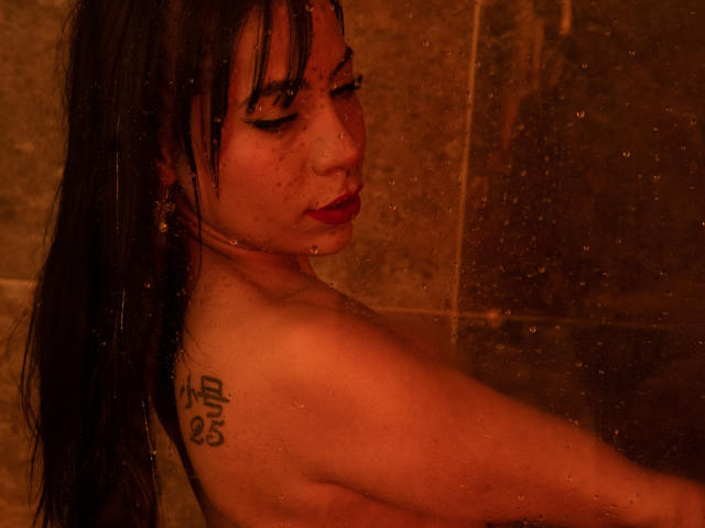 AmeliaRuso with wet black hair, red lipstick, and a tattoo on her right shoulder, standing in a brown-tiled shower with water droplets on her skin. Her eyes are closed.